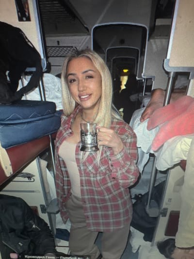 A blonde woman with light skin and dark eyebrows is shown in a cluttered interior that appears to be a train compartment. She is wearing a beige top and a red and grey plaid button-up shirt, and holds a metal cup holder with a glass in her right hand. Her hair is long and straight. The compartment contains bunk beds, luggage, and other personal items. The image appears to be a candid photograph, possibly taken on a mobile device, with a slightly grainy quality. The overall impression is that of a private, informal setting within a transportation vehicle. The city of Protasovo, Russia is not depicted in this image.