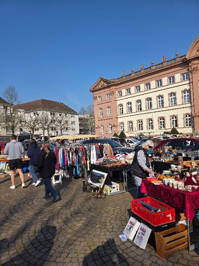 This image depicts an outdoor flea market in a public square within the city of Zweibrücken, Germany. The scene is dominated by a clear blue sky and brightly illuminated by daylight, casting distinct shadows across the paved ground, which features a dark, circular cobblestone pattern.

On the right side of the image, a prominent multi-story building with a reddish-brown facade, white-framed windows, and ornate architectural embellishments is visible. This structure is consistent with historical buildings in the Herzogvorstadt area or near Schlossplatz. In the background to the left, less ornate buildings with brown tiled roofs are discernible, alongside a few bare-branched trees, suggesting a late winter or early spring season.

The market features multiple vendor stalls. In the foreground on the right, a gray-haired woman wearing sunglasses, a striped shirt, and a dark vest stands behind a table draped in a red tablecloth, which displays numerous ceramics, mugs, and other small decorative items for sale. A red plastic crate containing blue objects, two folded newspapers, and a small wooden rack are positioned on the ground beside her stall. To her left, another stall presents racks overflowing with various articles of clothing, including shirts and jackets.

Several individuals are present, engaging with the market. Three people are walking in the mid-ground, primarily with their backs to the camera: a male in a gray hooded sweatshirt and dark shorts, followed by an individual in a dark blue jacket and jeans, and another in a dark jacket and blue jeans. Additional blurred figures are visible further within the market activity. Multiple vehicles, including SUVs and vans, are parked behind the stalls in the mid-to-background.