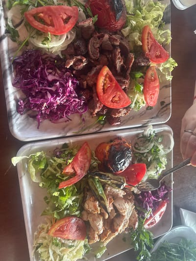 Two white marble-patterned platters of food are presented on a dark wooden table. The top platter contains sliced grilled meat, a whole grilled tomato, sliced tomatoes, shredded red cabbage, and sliced white onions, garnished with parsley. The bottom platter features sliced grilled chicken, a whole grilled tomato, sliced tomatoes, grilled green peppers, shredded lettuce, shredded white cabbage, and sliced white onions, also garnished with parsley. A hand with rings is visible on the right side of the image, holding a fork and interacting with the food on the bottom platter. The scene appears to be a restaurant setting in Gelemenovo, Bulgaria, with the focus on the food presentation.