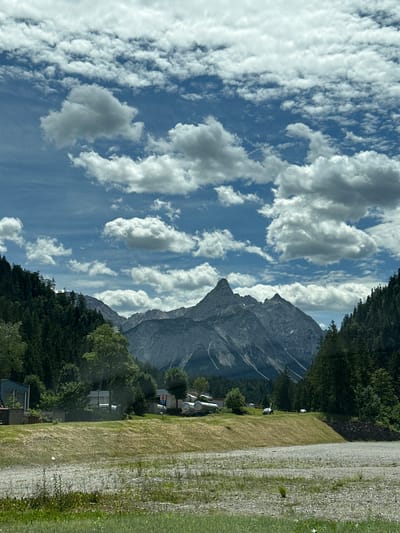 The image depicts a mountain landscape under a sky filled with cumulus clouds. In the foreground, a gravelly area with sparse vegetation leads to a grassy slope. Beyond the slope, there is a collection of industrial structures and vehicles. To the left, a blue building is visible, next to some cylindrical tanks. Further to the right, a small white building with a red sign and more tanks are present. Several vehicles, including cars and what appear to be tankers, are parked or driving on the area behind the grassy slope.

The middle ground is dominated by a dense forest of evergreen trees on either side of the frame, with the mountain range forming the backdrop. The mountains are rugged and rocky, with some patches of snow visible. A prominent, jagged peak stands out in the center of the mountain range.

The scene is likely located in the vicinity of Ehrwald, Austria, given the characteristic alpine scenery. The overall impression is of a rural or semi-industrial area situated at the foot of a majestic mountain range. The lighting suggests it is daytime, with the sun illuminating the clouds and the landscape, creating areas of light and shadow. No people are clearly identifiable in the image.