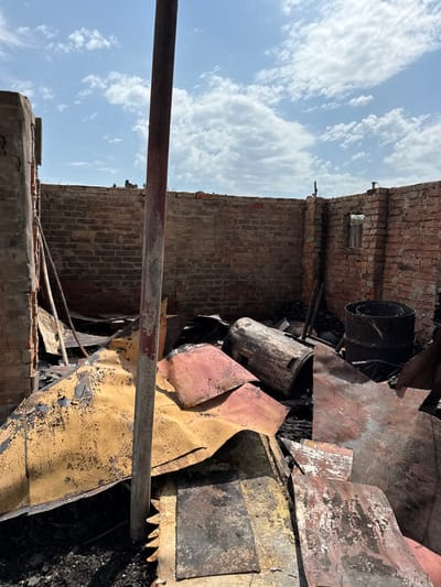 The image captures the interior of a severely damaged structure in Nedryhailiv, Ukraine, under a blue sky with scattered white clouds. The scene consists of standing red brick walls, which are the primary remaining elements of the building's enclosure, with an exposed upper edge indicating the absence of a roof. A small, rectangular opening, possibly a window frame, is visible in the far right wall. The ground is covered with a substantial accumulation of charred and warped metallic debris. This debris includes large, curved sheets displaying remnants of yellow, red, and grey coloration, mixed with blackened material. Amidst the debris on the right, a large, blackened cylindrical object lies horizontally, and another similar, smaller cylindrical object stands upright closer to the wall. Various smaller metallic rods are scattered within the debris pile. A prominent, reddish-brown vertical post, appearing to be structural, extends from the foreground to the upper portion of the frame, slightly off-center to the left. The base of this post features jagged, saw-like protrusions. No individuals are present in the depicted scene.