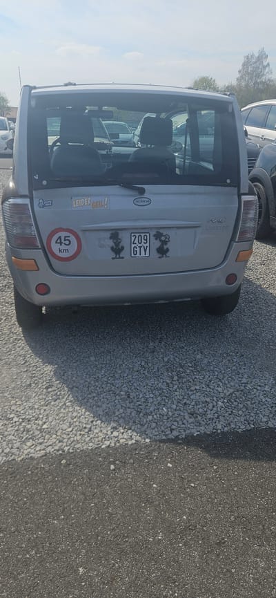 The image shows the rear of a silver Microcar MC2 Limited parked on a surface of gravel and asphalt. The car displays a license plate "209 GTY" and several stickers, including one indicating a maximum speed of 45 km/h, as well as cartoonish stickers and a sticker saying "Leider Geil!". The location is Delbrück, Germany. Other vehicles are visible in the background. No people are visible.
