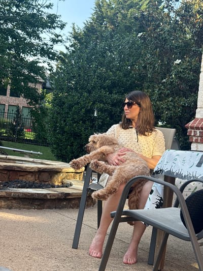 The image, taken in Southlake, Texas, USA, shows a woman sitting in a grey metal chair outdoors. She is wearing a light-colored, short-sleeved top, and sunglasses, and is holding a light brown Goldendoodle dog in her lap. The woman is barefoot. The chair is positioned on a concrete patio near a stone fire pit. The background includes a lush green tree and a glimpse of a brick building with dark window frames and a black metal fence. A table next to the woman holds items covered by a patterned cloth. The weather appears sunny.