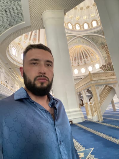 A man with dark hair and a beard takes a selfie in a large, ornate building with blue carpets and gold and white architectural details. The man is wearing a blue satin shirt with a repeating "GG" pattern. The background shows intricate Islamic architecture, including domes, arches, and decorative screens. The blue carpet is laid out in rows, typical of a prayer hall. The building appears to be the Akhmad Kadyrov Mosque, also known as "The Heart of Chechnya," located in Grozny, Chechnya, Russia. The city of Shali is not directly depicted or implied in this image.
