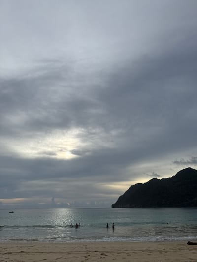 The image depicts a serene beach scene in Meunasahbale, Indonesia, likely during the late afternoon or early evening. The sky is filled with dramatic, swirling grey clouds, with patches of sunlight breaking through, suggesting the possibility of an impending sunset or a break in the weather. A dark, silhouetted mountain juts out from the right side of the frame, its peak covered in vegetation and its base meeting the water.
The calm, shallow ocean reflects the muted light of the sky. Several people are visible in the water, wading or swimming near the shore, their figures small against the vastness of the sea and sky. A small, dark boat is also present on the water further out. The sandy beach occupies the rightmost portion of the image, appearing relatively empty except for some subtle footprints and a piece of driftwood. The overall mood is one of tranquility and natural beauty, with the dramatic sky adding a touch of atmospheric intensity to the peaceful coastal setting. No text is visible in the image.