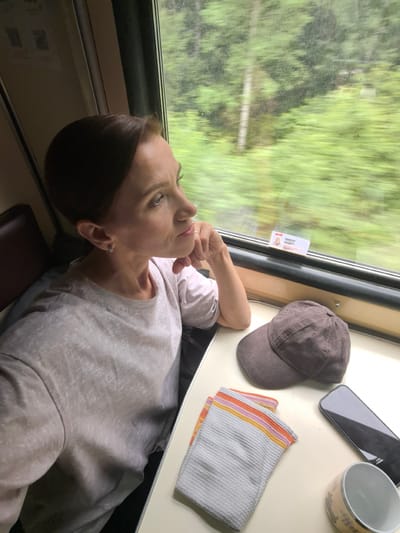 A woman with brown hair pulled back is sitting by a window on a train, looking out at the passing scenery. She is wearing a light gray t-shirt and has her hand resting on her chin, appearing contemplative. On the table in front of her are a brown baseball cap, a folded cloth with colorful stripes, a smartphone, and a mug. The train appears to be traveling through a rural area, with blurred green trees and vegetation visible outside the window. The setting is consistent with the city of Kuz'minka, Russia, given the context.