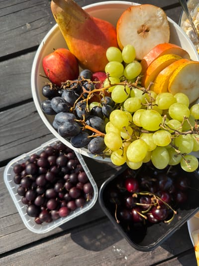 A close-up, high-angle shot depicts a fruit platter and two containers of berries on a weathered wooden surface, presumably outdoors. The wooden surface appears to be dark gray planks. In the center, a white bowl holds an assortment of fruits: a whole, large pear with red and yellow skin, sliced pears revealing their core and seeds, a red apple, green grapes, and dark purple grapes. To the left of the bowl is a clear plastic container filled with dark red berries, possibly blackcurrants or gooseberries. To the bottom right, a black container holds dark red cherries with their stems. The lighting suggests it is daytime, with sunlight casting shadows. The scene appears to be set in Kuliki, Russia, though this specific detail cannot be confirmed from visual cues alone. No people are visible in the frame.