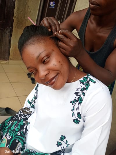 In Kuje, Nigeria, a woman is having her hair braided by another person. The woman receiving the braid is looking towards the camera with a slight smile, her head tilted to the side. She is wearing a white shirt with a black and green floral pattern on the sleeves and around the neckline. A pink comb is visible in her hair. The person braiding her hair is partially visible, wearing a dark gray tank top and their hands are focused on the braiding. The background appears to be an outdoor setting with tiled flooring and a doorway. The location is identified as Gui, Nigeria.