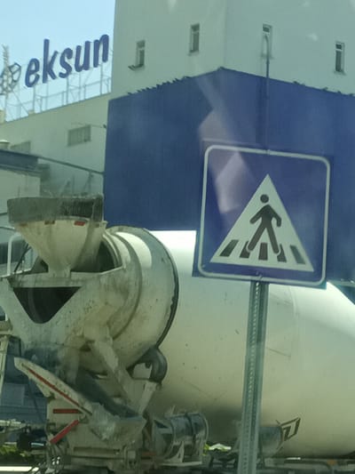 In the city of Konya, Turkey, a concrete mixer truck is parked in front of a white building with blue accents. A pedestrian crossing sign is visible in the foreground, positioned to the right of the truck. The sign features a blue square with a white triangle containing a black silhouette of a person walking across a crosswalk. The truck itself is partially obscured by the sign and the building. The building appears to be a commercial establishment with the word "eksun" displayed prominently on its upper facade in blue lettering. The sky is clear and bright, suggesting it is daytime. The overall scene depicts an urban industrial or commercial area.