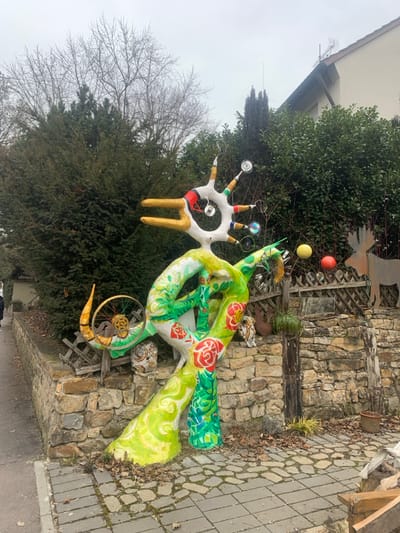 The image depicts an outdoor scene in Nürtingen, Germany, featuring a large, brightly colored, abstract sculpture positioned alongside a paved pedestrian walkway.

The primary subject is a multi-component sculpture situated on top of a low, dry-stacked stone retaining wall. The sculpture's upper section is predominantly white, featuring two golden, duck-bill-like prongs extending forward. A circular aperture is present in the center of this white section, from which multiple appendages radiate upwards and outwards, tipped with various small, colorful objects, including reflective discs and cylindrical forms. Red and black linear patterns are visible on this upper segment. The sculpture's main body comprises intertwined, organic forms painted in vibrant green and yellow hues, resembling stylized tree trunks or large vines. Several distinct red rose motifs are painted onto these green and yellow surfaces. Integrated into the mid-left portion of the sculpture is a bicycle wheel with a silver rim and yellow spokes, appearing as a decorative element.

Behind the stone wall and the sculpture, a dense, dark green evergreen hedge forms a significant backdrop. Above and behind this hedge, to the right, a section of a residential building with a brown sloped roof and a light-colored facade is partially visible. A thin antenna or lightning rod projects upwards from the roofline. To the right of the sculpture, behind the hedge, a rustic wooden fence is visible, featuring cut-out shapes, including a brown silhouette resembling a moose or deer, and decorative elements such as a yellow and a red spherical object.

The foreground consists of a pedestrian walkway paved with irregular grey pavers, many of which are square. Numerous dry, brown leaves are scattered across the pavement and around the base of the sculpture. To the far left in the background, a person is partially visible, walking away from the camera on the sidewalk, largely obscured by leafless deciduous trees and other vegetation. The sky is overcast and grey, indicating a non-sunny day. An empty terracotta pot is visible on the ground to the right of the sculpture.