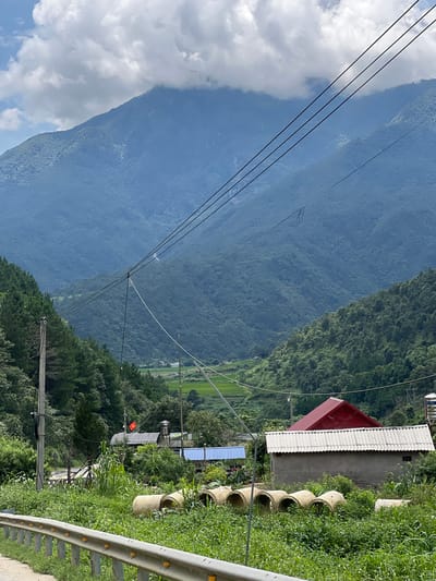 In Bình Lư, Vietnam, the image captures a mountainous landscape with a cloudy sky. In the foreground, a road barrier is visible with yellow reflectors. To the right of the road, there are several large concrete pipes lying in overgrown grass. Behind the pipes, a small building with a corrugated metal roof and a red-tiled roof is situated. Further back, a Vietnamese flag is flying above a structure. The background features lush green, tree-covered mountains that recede into the distance, with some sections obscured by clouds. Power lines stretch across the frame from the left to the right, passing in front of the mountains. A utility pole with wires is also present on the left side of the image. The overall scene depicts a rural, mountainous area.