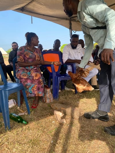An outdoor scene in Itobe, Nigeria, shows a group of individuals gathered under a light-colored canopy on dry, grassy ground. In the foreground, a man wearing a light shirt and dark trousers holds a live brown chicken with both hands, presenting it towards a seated woman. The woman, with dreadlocks and wearing a patterned dress, red sandals, and a gold necklace, sits on a blue plastic chair, her right arm slightly extended. A plaid tote bag rests beside her chair. Behind her, other individuals, predominantly men, are seated on blue plastic chairs, observing the central interaction. One man in a white shirt and beard is seated to the right of the woman, also looking towards the chicken. On the ground in the lower left, a green Heineken beer bottle lies on its side amidst some litter, including a broken white plastic container. In the lower right, a large, elongated yam or similar tuber lies on the ground, next to another piece of broken white plastic. The background shows clear blue sky and distant structures.