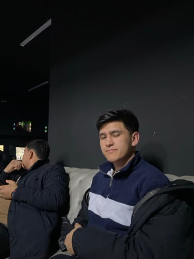 Two men are in a dark room. One reclines on a white cushion, his eyes closed, wearing a navy and white sweatshirt under a dark jacket. The other stands near monitors showing video game screens, wearing a dark jacket and light-colored shirt. The room has minimal lighting, with a black wall in the background. Long, thin lights illuminate the space on the left.