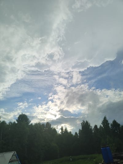 The image depicts a view of a cloudy sky over a dense line of trees. The sky is a mix of light gray and white clouds with patches of blue sky visible. The sun is partially obscured by the clouds, creating a diffused light. The trees in the foreground are dark green and appear to be a mix of deciduous and coniferous varieties. The bottom left of the image shows a partially constructed structure with a white roof and a blue tarp. In the bottom right, a large blue plastic barrel is visible, along with some other indistinct objects, possibly agricultural equipment. The overall scene suggests a rural or semi-rural environment. No people are visible in the image. The location is identified as Talitsa, Russia.