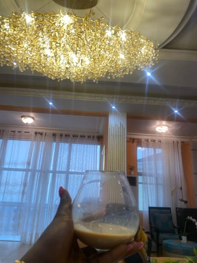 A person is holding a glass of liquid in their right hand. The liquid is a light brown color. The person is wearing a gold bracelet on their left wrist and has red nail polish on their right hand. The scene is inside a room with white curtains covering a large window in the background. There are several light fixtures in the ceiling. The room also contains an armchair and a small table. The scene is likely in a home in Buea, Cameroon, at the coordinates provided.