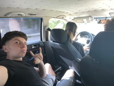The image shows the interior of a vehicle, likely a car, with four people inside. The viewpoint is from the back seat, looking towards the front. The image is rotated 90 degrees clockwise, making the people appear upside down.
In the foreground, the plush, tufted interior of the back seats is visible. A person's head with curly brown hair is visible in the lower left corner, facing towards a rear-view mirror. The mirror reflects a person's eyes.
In the middle ground, the back of the driver's seat and part of the passenger seat are visible. A person with long brown hair is seated in the back, facing forward. A person in the passenger seat is wearing a grey t-shirt and shorts.
In the background, the front of the car is visible. The driver, a young man with short brown hair and freckles, is in the driver's seat. He is wearing a black t-shirt and is making a "rock on" hand gesture. He is looking directly at the camera with a playful expression. The steering wheel and dashboard of the car are visible. The window shows a blurry view of trees and greenery, suggesting an outdoor setting, possibly a road trip. The lighting suggests it is daytime, likely a sunny day, given the bright light illuminating the interior.
There are no visible text elements in the image. The overall mood appears to be casual and friendly, with the subjects seemingly enjoying their time together. The location is Vlahovo, Bulgaria.