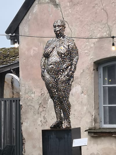 A statue of a full-figured person, constructed from numerous small, reflective pieces, stands on a dark pedestal. The statue is positioned in front of a textured, light-pink wall, which shows signs of age and wear. To the left of the statue, part of a building with a grey, tiled roof and a light fixture is visible, along with a grey metal fence. To the right of the statue, a light-blue window with multiple panes is set into the wall. A string of lights with visible bulbs runs horizontally across the upper portion of the scene. A small white sign is affixed to the pedestal in front of the statue. The statue is located in Kuldīga, Latvia.