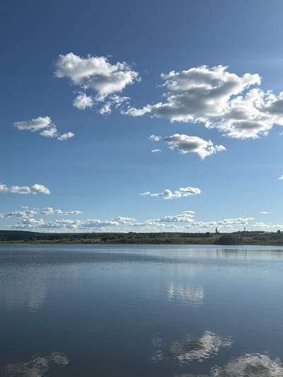 A wide, calm body of water stretches across the foreground of the image, reflecting the bright blue sky and scattered white clouds. The water is dark blue and appears still, with subtle ripples visible on its surface. In the middle ground, a dense line of green trees forms a natural barrier along the horizon. Beyond the trees, a faint line of hills can be seen, with a few distant industrial structures, possibly part of Taskino, Russia, silhouetted against the sky. The upper portion of the image is dominated by a vast blue sky filled with fluffy white cumulus clouds, some catching the sunlight and appearing brighter than others. The overall scene is serene and natural, with a hint of human presence in the distance.