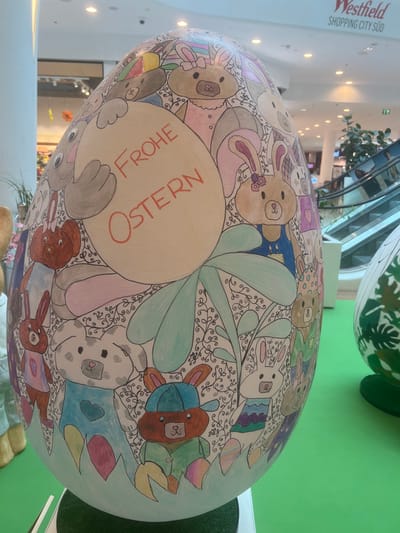 The image displays a large, anthropomorphic Easter egg prominently featured within the multi-level interior of the Westfield Shopping City Süd in Vösendorf, Austria. The egg, which dominates the foreground and midground, is decorated with numerous cartoon-style bunnies and other animal figures in various pastel colors (pinks, blues, browns, greens, grays) on a white base, interspersed with black swirly vine patterns and larger green leaves. A central white oval on the egg contains the red German text "FROHE OSTERN" (Happy Easter).
In the background, the modern architecture of the shopping mall is visible, characterized by white walls, multiple floors, glass railings, and an escalator descending on the right. Overhead, recessed lighting illuminates the space. A sign for "Westfield SHOPPING CITY SÜD" is visible in the upper right. The egg itself rests on a dark green circular base, and the surrounding floor area is covered with a bright, uniform green surface. A second, partially visible Easter egg, decorated with green and white patterns, is situated to the right of the main subject. No specific individuals are clearly identifiable, though distant figures suggest general mall activity.