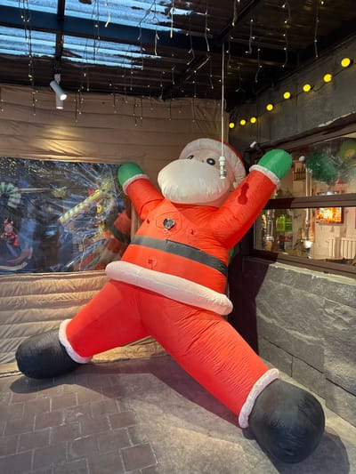 An inflatable Santa Claus figure is positioned prominently in a decorated indoor or enclosed outdoor space within Lishan District, China. The Santa, predominantly red with white trim, a black belt, and green gloves, stands upright with arms raised and spread. A dark circular port is visible on its chest. Its face is white with a beard and a white hat. A thin silver rod hangs from the ceiling in front of its head.

The immediate environment features a patterned reddish-brown brick floor on the left, transitioning to a smooth dark grey surface on the right. The ceiling is dark, possibly corrugated, and adorned with numerous strands of hanging white "icicle" style fairy lights. A white track light with two spotlights is mounted on the left side of the ceiling.

To the left of the Santa, a window-like opening is covered by a clear plastic sheet, behind which blurred holiday decorations, including a striped candy cane, are visible. Below this, a horizontally ribbed, light-colored blind or wall panel is present. To the right, a dark grey, textured stone wall contains a horizontal window revealing bottles and items on shelves, suggesting an interior space like a bar or restaurant. Above this window, a string of round yellow lights illuminates the wall. No individuals are present in the scene.