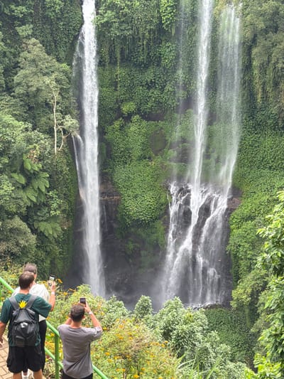 A tall, multi-tiered waterfall surrounded by lush, green vegetation cascades down a rocky cliff face. A group of tourists stand on a paved viewing platform with a metal railing, capturing the scene with their mobile phones. One person wears a backpack with a bright yellow flower attached. The surrounding landscape is verdant, with trees and shrubs adding to the natural beauty of the location.