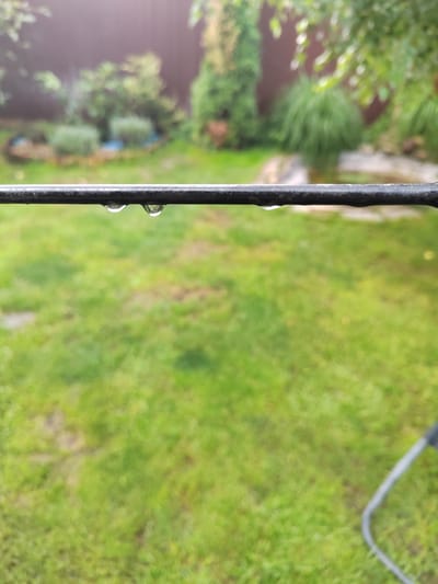 This media file depicts a close-up of a dark, horizontal metal bar with two water droplets clinging to its underside. The droplets are clear and refract light, indicating recent rainfall. The bar is the primary subject in the foreground, with the background significantly blurred to create a shallow depth of field.

The background reveals a lush green lawn, suggesting a garden or backyard setting. Behind the lawn, a wooden fence is visible, bordering the property. Further back, a variety of green foliage, including bushes and trees, are present, some appearing manicured and others more natural. The overall impression of the background is that of a well-maintained outdoor space.

The lighting suggests it is daytime, possibly overcast due to the presence of water droplets and the diffused light. The soft, diffused light and the water droplets strongly indicate that it is raining or has recently rained. The location is identified as Yuzhnyy, Russia, which provides a geographical context for the outdoor scene. No people or specific activities are depicted. The image evokes a sense of tranquility and the quiet beauty of nature after a rain shower. There is no visible text within the image.