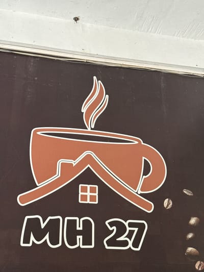 The image displays a sign with a coffee cup logo and the text "MH 27". The logo is brown and white, featuring a steaming coffee cup with a house silhouette inside it. The text "MH 27" is also in white with a brown outline. The sign is affixed to a dark brown background, with scattered coffee beans visible on the right side. Above the sign, a white, textured surface is visible, with a small dark object on it. The sign appears to be an exterior sign, likely for a business. The context provided states this is located in Meunasahmanyet, Indonesia, however, no specific environmental cues within the image confirm this location. There are no people or animals present in the image.