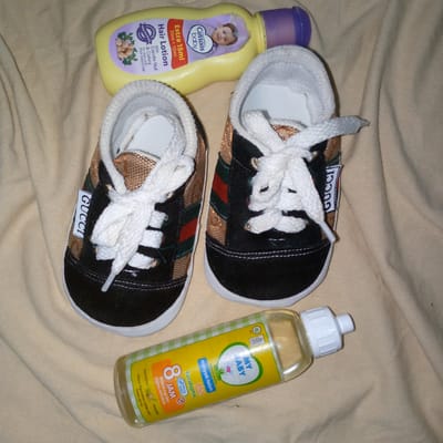 A flat lay photograph displays a collection of baby-related items on a wrinkled, light beige fabric surface. In the upper part of the frame, a yellow bottle of "Cussons baby Hair Lotion" with a purple screw cap is oriented horizontally, angled slightly towards the upper right. The label indicates "Extra 15ml 35ml + 15ml" and features text specifying "Dermatologically Mild Formula Tested" and "With Candle Nut & Celery for dark and thick baby's hair," alongside an image of a baby's face. Centered below the lotion is a pair of black and brown baby shoes, positioned side-by-side with their toes pointing towards the top of the image. The shoes feature white rubber soles and white laces, which are tied. The main body of the shoes is black with a patterned brown/gold fabric section on the sides, accented by distinct red and green stripes. A white tag, vertically displaying "GUCCI" in black text, is attached to the outer side of the left shoe. In the lower portion of the image, a clear plastic bottle of "MY BABY minyak telon plus" with a white screw cap is placed horizontally, angled slightly towards the upper left. Its yellow label includes a graphic of a baby, text stating "8 JAM" (8 hours) in red, and mentions "Eucalyptus" and "Citronella," with additional text in Indonesian indicating "Melindungi dari gigitan NYAMUK" (Protects from mosquito bites) and "Netto 60 ml." No individuals or actions are present. The visual content does not provide information to confirm the location as Meunasahblang, Indonesia.