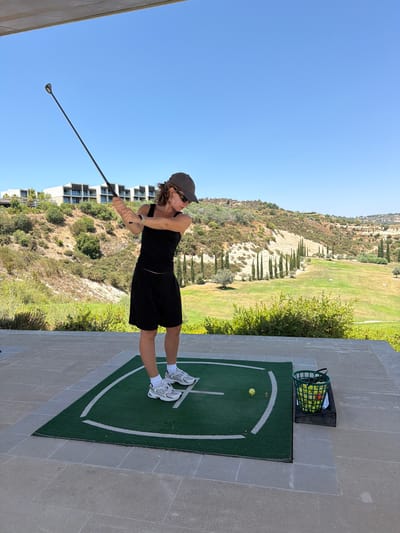 The image depicts a woman practicing golf on a driving range in Tsada, Cyprus. She is wearing a black tank top, black shorts, a grey baseball cap, sunglasses, and white athletic shoes. Her body is positioned as if she is in the middle of a golf swing, with her arms extended and holding a golf club. She is standing on a green mat with white markings, which is placed on a paved surface. To her right, there is a green basket filled with yellow golf balls.

The background features a lush green golf course with cypress trees and rolling hills dotted with vegetation. In the distance, modern buildings are visible on the hillside. The sky is clear and blue, indicating a sunny day. The overall scene suggests an outdoor recreational activity in a scenic location.