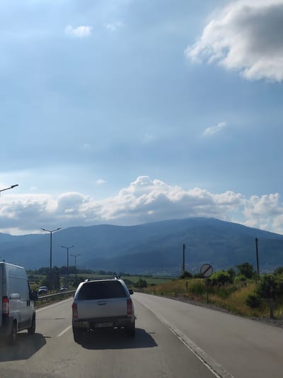 The image depicts a view from a vehicle traveling on a highway. In the foreground, a silver pickup truck with a camper shell is visible, displaying the license plate "BT 1930 KT". To the left, the rear of a white van is partially visible. The road ahead curves to the right, with a speed limit sign indicating the end of any restrictions. The landscape features rolling green hills and mountains in the background, with scattered trees and vegetation along the roadside. The sky is a light blue with scattered white and gray clouds. Streetlights are visible along the highway, suggesting it is a developed area. The presence of the license plate suggests a location in Bulgaria. Based on the scenery and the specific license plate format, the location is likely near Golemo Buchino, Bulgaria.