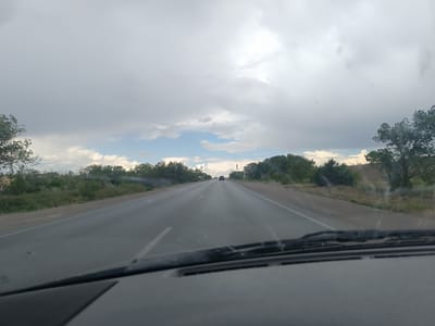 The image shows a view from inside a vehicle driving on a highway. The road ahead curves slightly to the right. There are trees and vegetation on both sides of the road, with hills in the background. The sky is overcast with dramatic clouds. There are a few other vehicles visible on the road in the distance. The dashboard and windshield wipers of the vehicle are visible in the foreground. The scene appears to be set in a rural or semi-rural area.