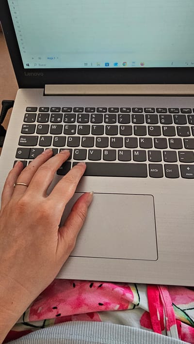 This is a close-up, top-down shot of a person's hand resting on a laptop keyboard. The laptop is open, with a spreadsheet visible on the left screen and the keyboard on the right. The keyboard is a standard QWERTY layout with Spanish characters, indicated by keys like "Ñ" and "Ç". The hand, positioned over the "C", "V", and "B" keys, appears to be in a resting position, suggesting the person is either pausing from typing or about to begin. A thin gold ring with a small stone is visible on the ring finger.

The laptop itself is silver. To the right of the laptop, a colorful, patterned fabric is visible, featuring large pink and green fruit motifs that resemble watermelons. The setting appears to be indoors, with ambient lighting, and no clear indicators of the time of day or weather.

The screen on the left shows a spreadsheet with numbered rows and columns, and the text "Hoja 1" and "Buscar" are visible. The laptop branding "Lenovo" is clearly visible on the lower bezel. The presence of a spreadsheet and the laptop suggest an activity related to work, study, or personal organization. The cropped nature of the image focuses attention on the interaction between the hand and the keyboard, hinting at a moment of focused activity or contemplation. The overall mood is neutral and functional, highlighting the tools of digital engagement.