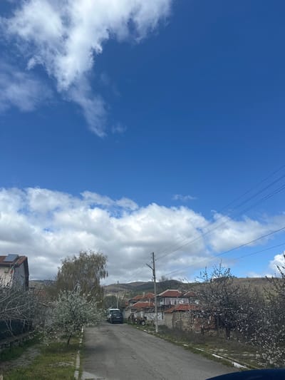 The image depicts a street scene in Gavrailovo, Bulgaria. The sky occupies the left half of the frame, showing a clear blue expanse interspersed with white cumulus clouds. Electrical power lines are visible stretching across the sky.
On the right side of the frame, a paved road runs through a residential area. Several houses with red tile roofs are visible. A black vehicle is parked along the side of the road. Trees in bloom line the roadside, with blossoms suggesting springtime. A tree in front of the house also has white blossoms. The terrain appears to be slightly hilly, with a hill in the background.