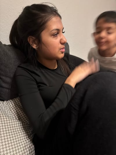 The scene is an interior setting in Tuttlingen, Germany. A young adult female with dark hair secured in a ponytail is positioned in the left foreground, wearing a black long-sleeved top. Her head is turned to her right, and her right arm is raised, with the hand showing motion blur. A child, heavily motion-blurred and wearing a light-colored top, is visible in the right background, facing the female. The child's facial features are indistinct due to motion. The female rests against a dark gray cushion. A gray and white houndstooth-patterned cushion is to her immediate left. The background wall is plain and light-colored.