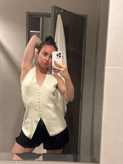 A young woman is taking a mirror selfie in what appears to be a bathroom. She is holding an iPhone in her right hand, and her left arm is bent with her hand near the back of her head. She is wearing a light-colored, button-down vest and black shorts. Her hair is pulled back into a bun. The bathroom has grey and white tiles. A partial reflection shows a shelf above the sink area, along with a towel and a dark-colored cabinet. The image does not provide enough information to confirm if it was taken in Baan Klong Prao, Thailand.