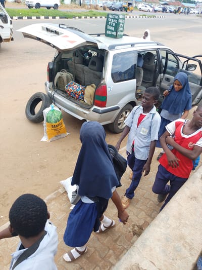 This media file captures a scene at Oja Oba, Oko Erin, Nigeria, likely during daylight hours. The central focus is a silver minivan with its tailgate open, packed with luggage. A tire and a large bag of rice labeled "BANYAN RIZ PARBONED 5%" are placed beside the vehicle on the dusty ground.
Several individuals, appearing to be students, are interacting around the minivan. Two young boys, one in a striped shirt and blue pants, and another in a red vest and blue pants, are standing near the open passenger door. A young girl in a blue hijab is also near the passenger door, seemingly assisting or interacting with the boys. Another figure, also in a dark blue hijab, is seen from behind, wearing a blue skirt and white socks with sandals. The expressions on the faces of the visible individuals suggest a moment of preparation or departure, possibly for a trip or journey.
In the background, a road with other vehicles, including a tricycle and cars, is visible. A sign with text, partially legible as "ISEYIN OKEHO OTU SAKI IGBO ORA," is placed on the roof of the minivan. The overall setting suggests a busy transport hub or a roadside stop in a Nigerian community.