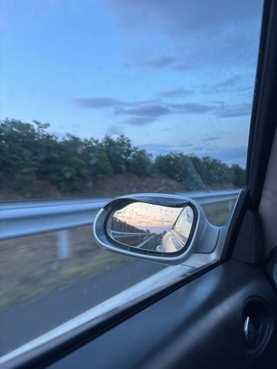 The image depicts a view from inside a car, looking out the side window and into the side-view mirror. It is a cloudy day with a pale blue sky, and there are raindrops visible on the car window. The car appears to be on a highway, with a metal guardrail running alongside the road and a line of trees on a hillside in the background. The side-view mirror reflects the highway stretching into the distance, with the sky and clouds visible in the reflection. The interior of the car, specifically the door panel and a speaker grille, is visible in the foreground on the right side of the image. There is no indication of the specific location being Zheleznik, Bulgaria; the image provides no discernible geographic markers to confirm this.