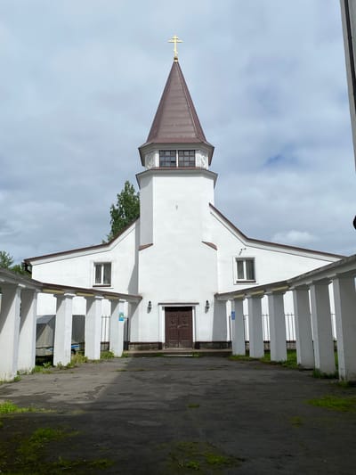 A frontal, slightly low-angle, eye-level shot captures the exterior of a white church in Sortavala, Russia. The church features a prominent, brown, spire-like roof topped with a golden cross. A rectangular section below the spire has a band of windows. The main body of the church is white with a brown-trimmed, steeply pitched roofline. There are two rectangular windows on the main facade, one on each side, and a set of double brown doors in the center. Flanking the entrance is a white colonnade with square pillars and a brown roof extending outwards. The ground in front of the church is paved with dark asphalt, with patches of overgrown grass. To the left of the entrance, behind the colonnade, a covered trailer is visible. The sky is overcast with visible clouds.