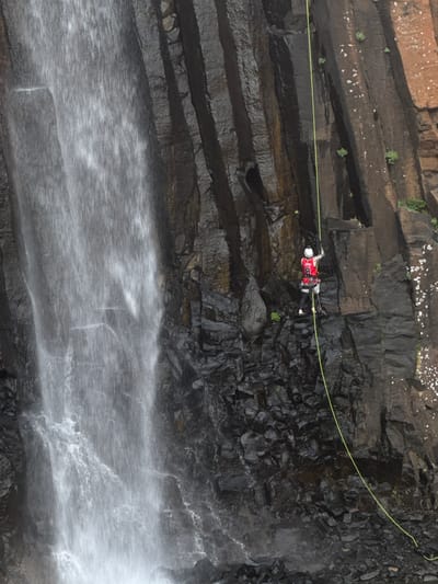 In a scene captured from above, a person rappels down a steep, layered rock face. The individual is clad in a red and grey outfit, complete with a white helmet and safety gear, suspended by a bright yellow rope that stretches diagonally across the frame. The rock formation is characterized by dark, wet layers, with streaks of lighter brown or tan rock interspersed. Small patches of green vegetation cling to the higher sections of the cliff. A significant waterfall cascades down the lower portion of the rock face, creating a misty spray that obscures the base of the cliff and blends into the rocks below. The water's movement is captured with a slight motion blur, indicating a long exposure or fast-flowing water. The lighting suggests it is daytime, possibly overcast, as there are no strong shadows. The overall impression is one of an extreme outdoor adventure activity taking place in a rugged, natural environment.