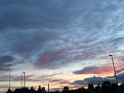 This image depicts a cloudy sky over Laagri alevik, Estonia, likely during sunset or sunrise. The sky exhibits a gradient of colors, with shades of blue, gray, pink, and orange visible in the clouds. Several tall, modern streetlights are positioned along the lower part of the frame, silhouetted against the sky. In the background, the dark silhouettes of trees and some buildings can be observed. A parking sign with a blue 'P' symbol is also visible to the right of the frame. The scene appears to be an outdoor urban or suburban environment.