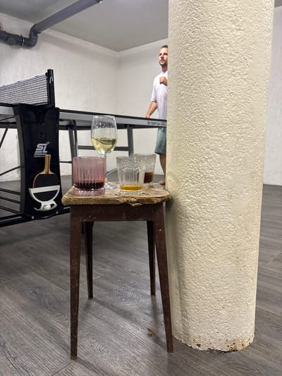 In a room with gray wood-patterned flooring and white walls, a man in a white t-shirt and blue shorts stands behind a ping pong table, his left arm raised. In the foreground, a small, rustic wooden stool with metal legs holds three glasses. The leftmost glass is pink and filled with a dark liquid. Next to it, a clear glass contains a light amber liquid. To its right, another clear glass holds a dark liquid. A white wine glass with a light yellow liquid is positioned behind the pink glass. The ping pong table has a black net and a black paddle with red and white accents is clipped to its side. A large, textured, off-white pillar is situated to the right of the stool, partially obscuring the man. A gray pipe runs along the ceiling. The scene appears to be indoors, possibly in a recreational area. The location within Лугаваслабадскі сельскі Савет, Belarus is not discernible from the visual information.