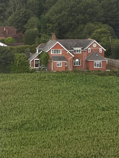 The image displays a large, detached, red-brick residential property situated at the edge of a significant green field, likely cultivated, in the city of Exeter, United Kingdom. The multi-story house features a dark grey slate or tile roof with multiple gables and dormer windows, white window frames, and numerous multi-pane windows of varying sizes, including a prominent circular window (oculus) on the upper-right facade. Several chimneys are visible on the roofline. To the immediate left of the house, a small, transparent lean-to structure, possibly a greenhouse, stands near the entrance, with white flowering plants visible beside it. A wooden post-and-rail fence runs along the right side of the property, bordering an unpaved track. The background is dominated by a dense, dark green treeline, indicating a wooded hillside, with glimpses of other residential building rooftops and hedges visible to the left of the main house. No individuals are present in the scene. Natural daylight illuminates the entire setting uniformly, suggesting an overcast or ambient condition.
