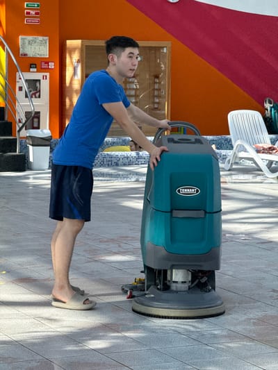 A young man is operating a Tennant brand floor scrubber on a tiled floor. He is wearing a blue t-shirt, dark blue shorts, and tan slip-on sandals. The background includes a tiled pool area with a white lounge chair, a red and orange wall, and fire safety equipment mounted on the wall. The location is in Kogalym, Russia.