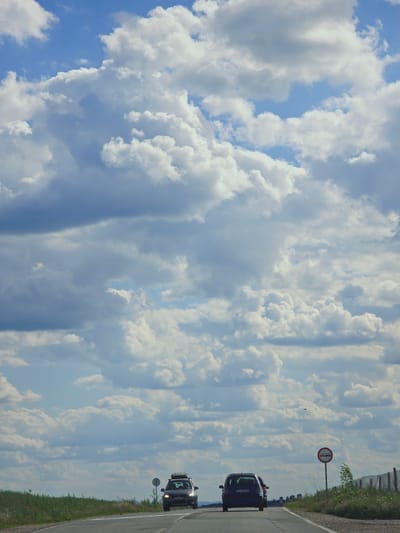 The image depicts a rural road scene in Kichevo, Bulgaria, under a cloudy sky. Two cars are parked on the roadside. A road sign indicating "no overtaking" is visible. The landscape consists of a road, roadside grass, and a background filled with a sky full of clouds.