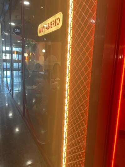 The image displays the exterior of a commercial establishment located in Leganés, Spain, viewed through a large, reflective glass facade with a red frame. An internally illuminated sign, reading "MUY ABIERTO" in red capital letters on a white background, is mounted above the interior. To the right of the glass facade, a vertical panel is backlit with orange light, presenting a diamond grid pattern. Adjacent to this, a red vertical structure contains a bright, narrow, vertical red light strip. The overall illumination is warm, dominated by artificial orange and red light sources. The glass reflects a polished floor and architectural elements of an indoor hallway or concourse, including a mirrored, partially legible sign indicating "WOK center." Inside, at least three individuals are partially visible, seated at tables. One individual with dark hair is positioned on the right side of the interior, facing left, while another individual in a light-colored top is seated further left with their back partially to the viewer. No specific actions or interactions among the individuals are clearly discernible. The interior decor includes geometric wall patterns, a hanging plant, and recessed ceiling lights.