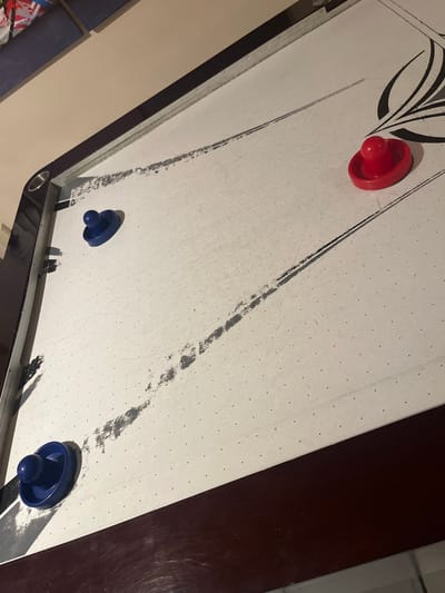 The image shows an air hockey table from an angled perspective. A red puck sits at the upper left, and two blue pucks are at the lower right and center. The table surface is white with numerous small holes and black lines marking the playing area. The lines have grey smudges along them. The table's frame is visible, with a dark wood-grain side at the right. A wall is in the background on the left.