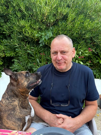 The image shows a medium shot of a man and a dog sitting outdoors, with a dense green hedge in the background. The man, who appears to be in his late 40s or 50s, has a fair complexion, short grey hair, and is wearing a dark blue t-shirt and grey shorts. He is looking directly at the camera with a slight smile. A pair of glasses are hanging from the neckline of his t-shirt. The dog, a brindle-colored breed with a white chest, is sitting to the left of the man and looking upwards and to the man's left. The dog's mouth is slightly open, showing its teeth. They are seated outdoors, possibly in a garden or patio area, as suggested by the gravelly ground visible in the lower right corner. The lighting appears to be natural daylight. There is no specific information within the image to indicate the location being Općina Baška, Croatia.