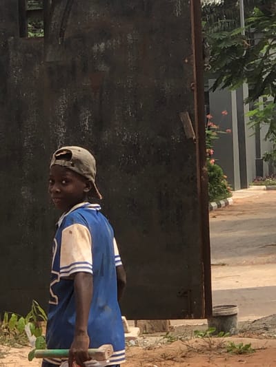 The scene, located in Abatete, Nigeria, depicts a young male subject in the mid-ground, slightly left of center. The individual, appearing to be a child, has dark skin and wears a light brown baseball cap reversed. He is dressed in a blue short-sleeved shirt with white sleeves and blue-and-white striped cuffs and hem, displaying noticeable dirt or smudges on the fabric. The subject is turned partially away but looks over his left shoulder directly at the viewer, exhibiting a slight smile. He holds a green-handled tool, possibly a trowel, in his right hand, which hangs at his side. The foreground consists of reddish-brown dirt. Behind the subject and to his left stands a large, dark, weathered metal gate or wall. To the right of this gate, an opening reveals a paved area with a black-and-white striped curb. Beyond this, green foliage, including trees and bushes with some orange flowering plants, is visible, along with a portion of a modern building featuring grey walls in the background. A white bucket or container rests on the ground near the bottom right edge of the gate.