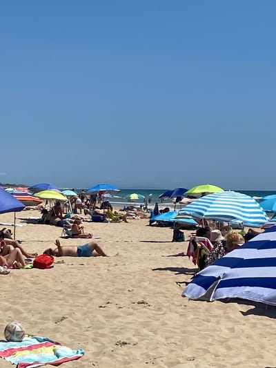 The image captures a sunny day at a crowded beach in Conil De La Frontera, Spain. The sandy shore is filled with people relaxing under numerous colorful beach umbrellas. In the foreground, a person lies on their back on the sand, wearing blue shorts, with their left arm raised. A red bag is situated near them. To the left and in the foreground, a striped beach towel with a soccer ball rests on the sand. Beachgoers are scattered throughout the scene, some seated, some lying down, and others are partially visible in the background, with the ocean and its gentle waves visible in the distance under a clear blue sky.