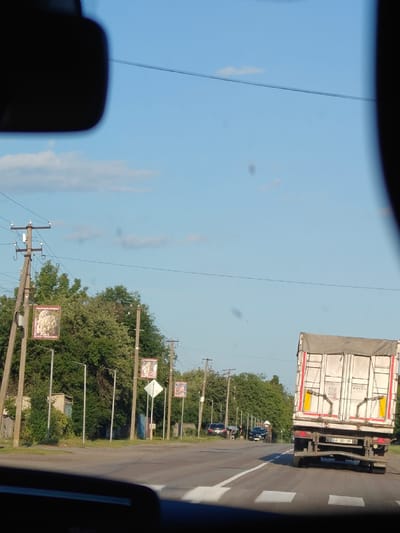 This media file captures a view from a vehicle, looking out at a roadside scene in Opishnia, Ukraine. The time of day appears to be late afternoon or early evening, as indicated by the warm, soft lighting and the clear blue sky with scattered clouds. The weather seems pleasant, suitable for outdoor activities.

The foreground is dominated by the interior of the vehicle, with a blurred rearview mirror and dashboard visible. The main subject of the image is the road ahead, with a white lane marker indicating the direction of travel. On the right side of the road, a large, white semi-trailer truck with red and yellow accents is prominently featured, moving away from the viewer. The truck has "BODEX" written on its side and a Ukrainian license plate reading "AA 1201 XF".

To the left of the road, a line of utility poles with electrical wires stretches into the distance. Several of these poles are adorned with rectangular posters or banners, some featuring images and text. One visible banner appears to be advertising the "MUSEUM OF POTTERY" in Opishnia, with text in Ukrainian. Further down the road, more poles with similar banners and streetlights are visible.

Behind the utility poles, lush green trees and dense foliage line the side of the road, suggesting a semi-rural or suburban setting. A few smaller buildings and a wooden fence are partially visible amongst the trees. In the distance, other vehicles are seen on the road, including cars and potentially other trucks. Two figures are standing near parked cars on the left side of the road, though their activity is unclear.

The overall impression is one of a typical day in a Ukrainian town, with everyday traffic and local advertisements on display. The scene is peaceful and ordinary, without any dramatic events or strong emotional cues.